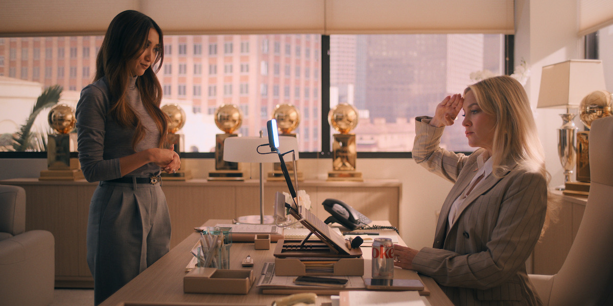 Two women in business attire are in a modern office with large windows and several gold awards on display. One woman stands while the other sits at a desk, saluting, with office supplies and a phone in view.