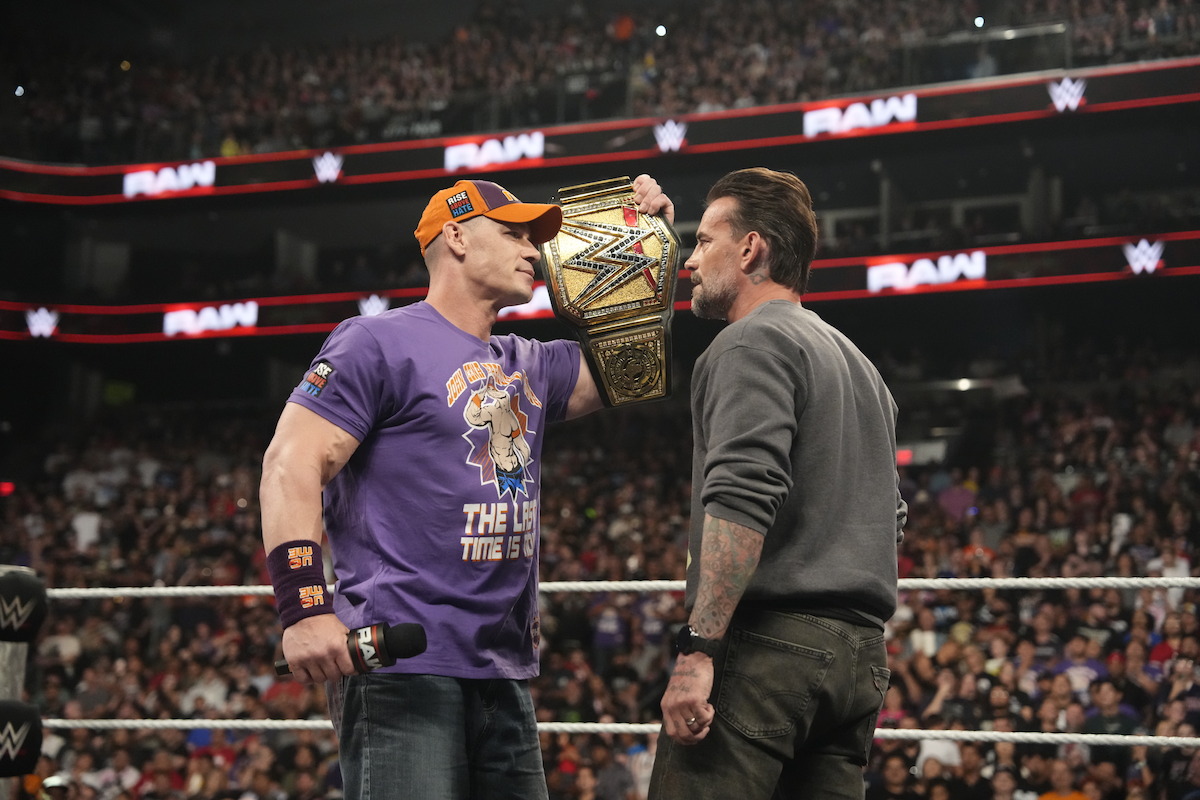 John Cena and CM Punk in a WWE ring face off, one holding a championship belt up to the other's face as a large audience watches in an arena setting.