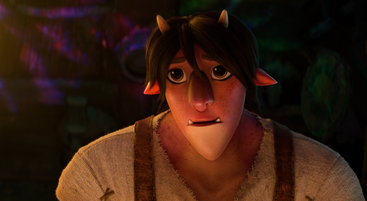 Animated character with small horns, pointy ears, and a concerned expression, wearing a rough shirt and suspenders, in a dimly lit, rustic environment with colorful light reflections on the wall behind.