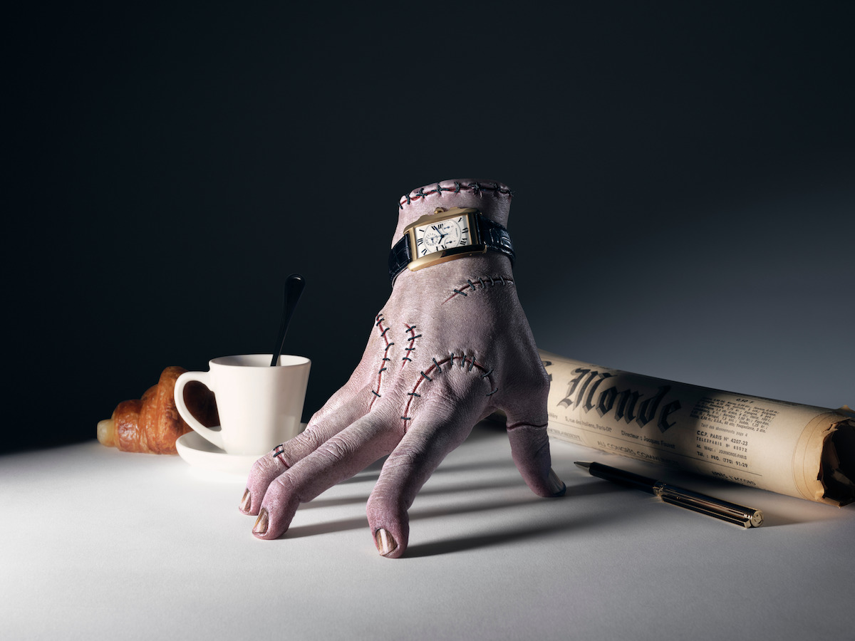 Thing wearing an analog wristwatch beside a coffee cup, croissant and newspaper.