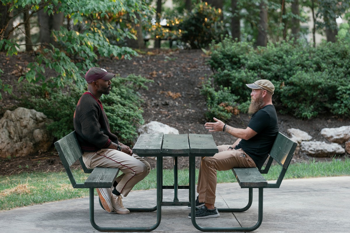 Two men sit across from each other talking at a green picnic table in a park. Trees, bushes, and rocks surround them, and the setting is calm and casual.