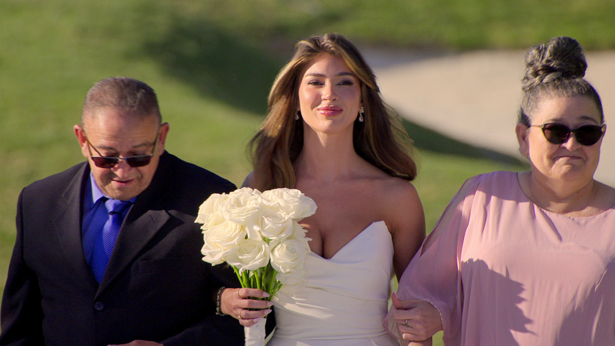 The bride holding white roses walks outside between an older man in a suit and an older woman in pink, all eager and smiling. The atmosphere is joyful, with a bright and sunny setting on green grass.