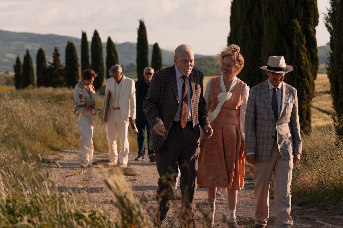 A group of elderly people in formal attire walk along a sunlit country path lined with tall trees, surrounded by grassy fields and hills, sharing a warm and relaxed moment together.