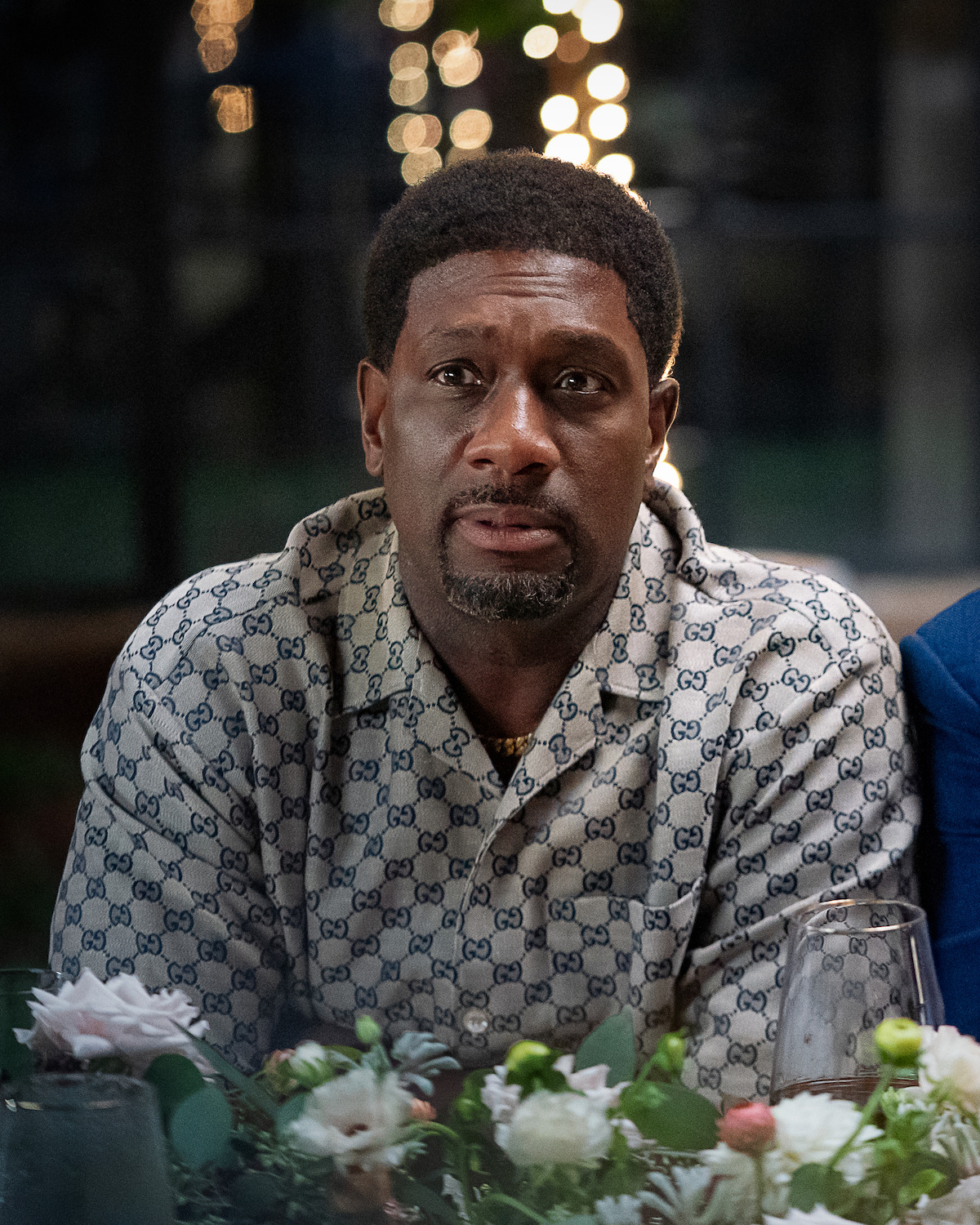 Man sitting at a decorated dinner table with floral arrangements, wearing a patterned shirt, indoors at night with string lights blurred in the background.