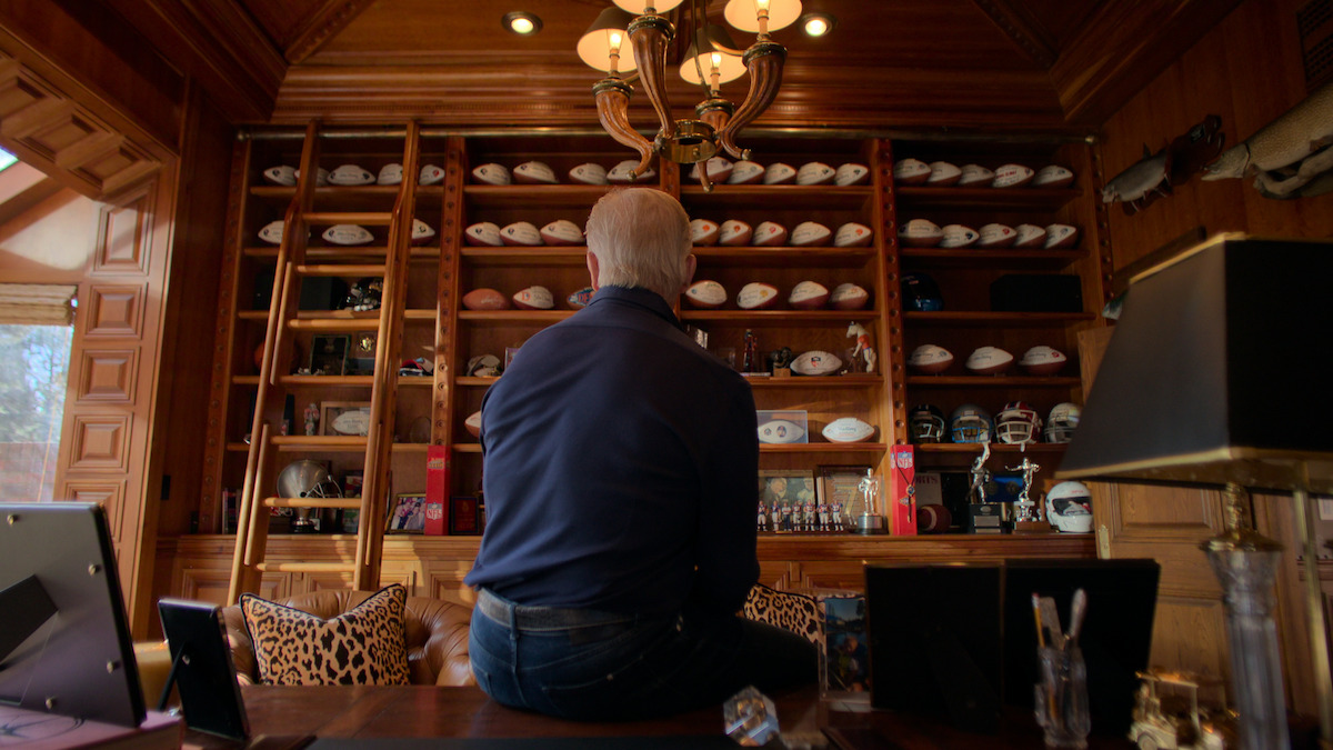 John Elway sits on a desk in a wood-paneled room facing shelves filled with football helmets, signed footballs, and trophies, with a ladder and animal mounts also visible in a well-lit study or office environment.