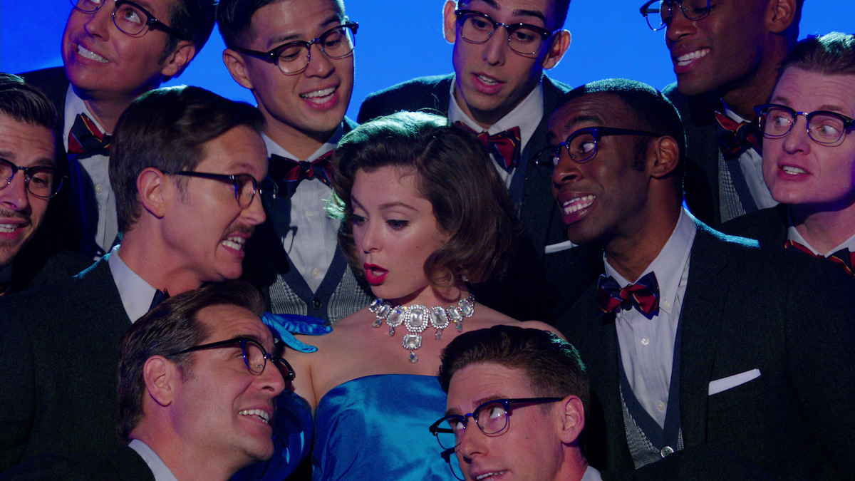 a woman in a blue dress sings while several men lean in close and sing along