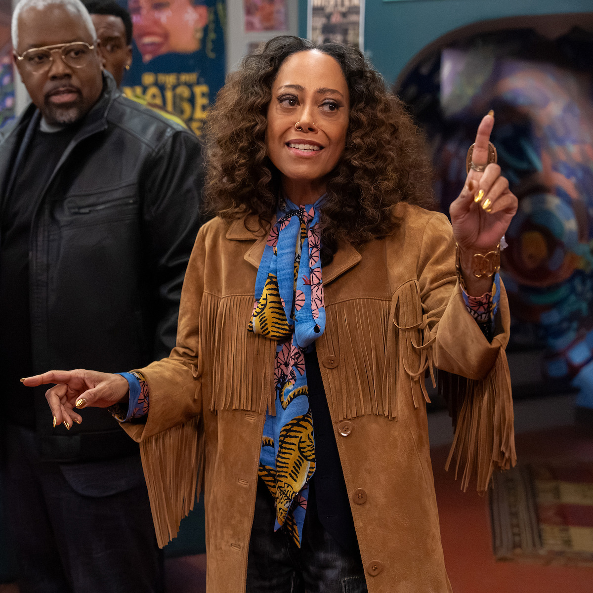 Woman with curly hair wearing a tan fringed jacket and colorful scarf gestures with both hands indoors, with a man in a leather jacket and posters visible in the lively background.