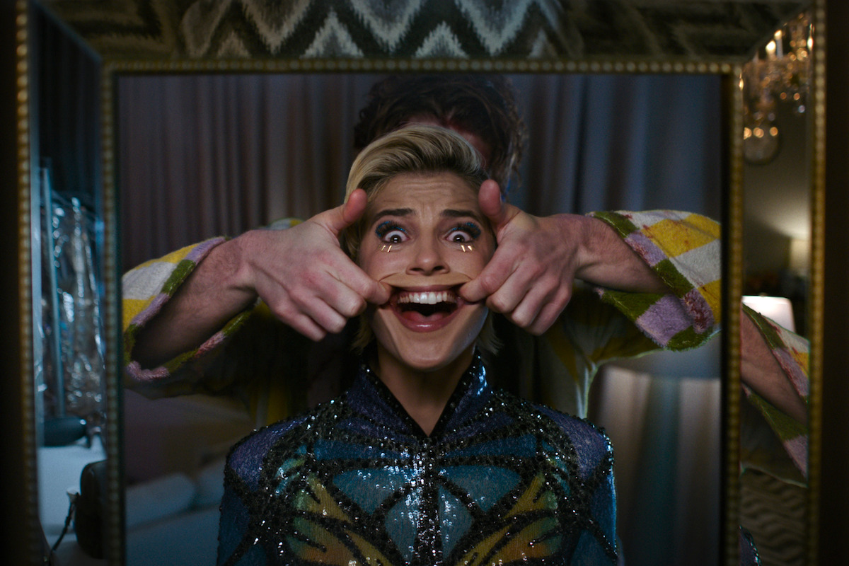 Person in sequined outfit smiling in a mirror as another person, partially visible, stands behind them stretching their mouth into a grin; indoor setting with curtains, soft lighting, and patterned decor in the background.