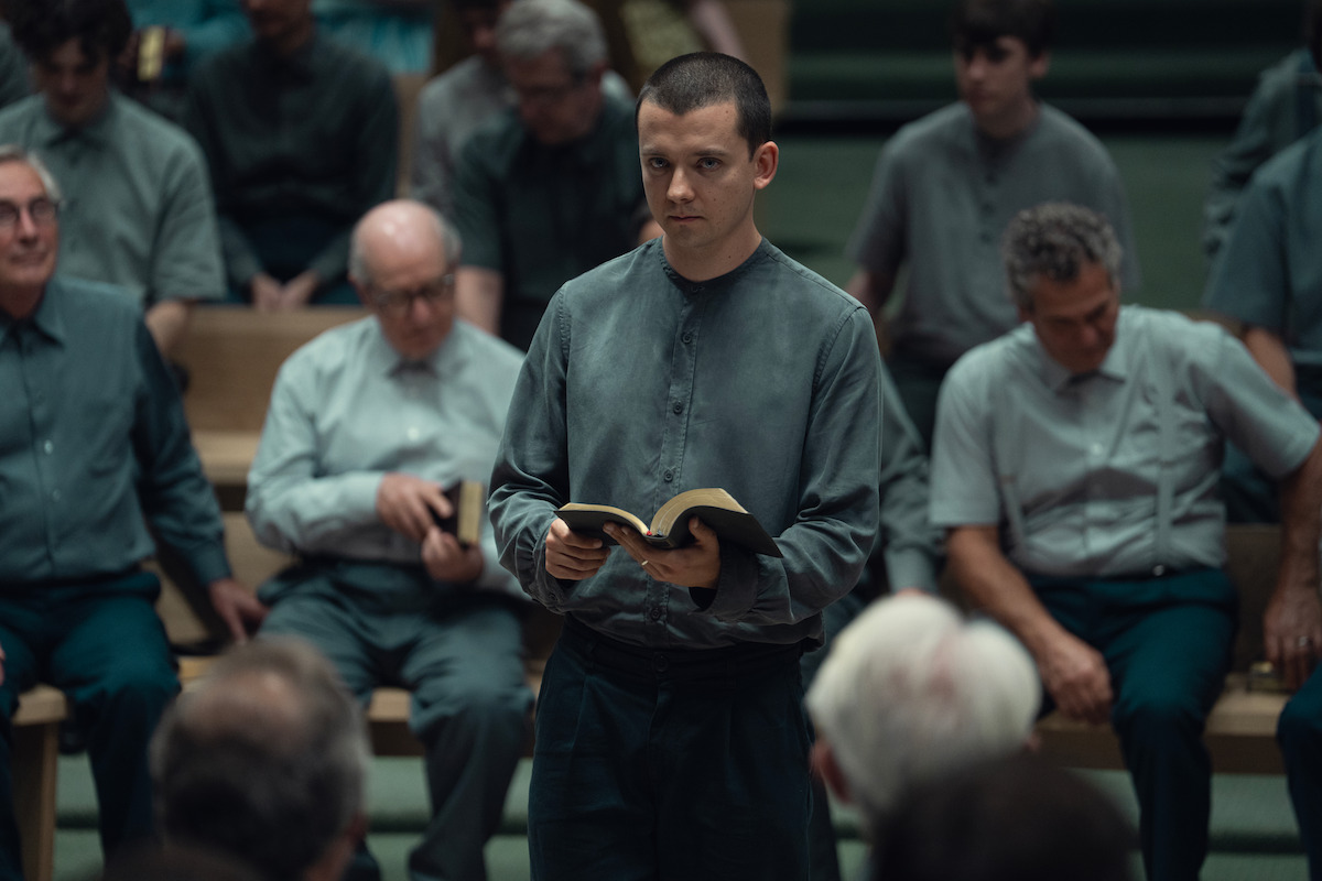 A young man stands holding an open book in a dimly lit room, surrounded by seated men in similar grey clothing, suggesting a formal or religious gathering, with attentive and solemn expressions.