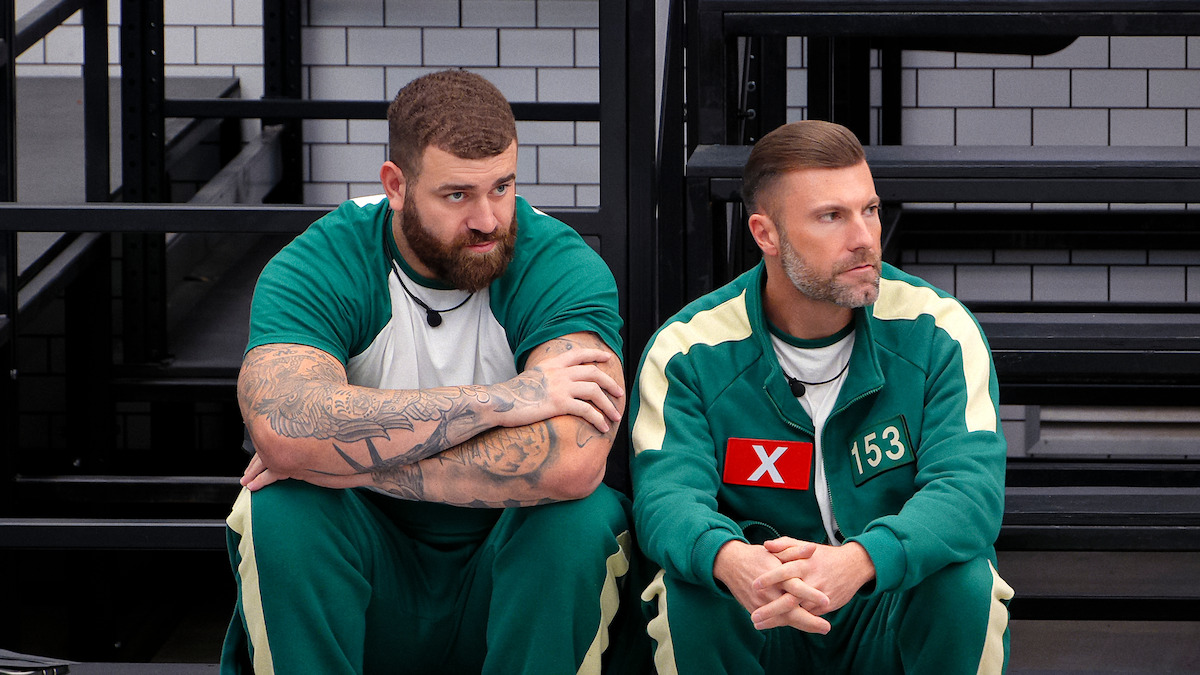 Two men in green tracksuits with numbers sit on a bench in a stark, tiled room, appearing pensive and tense, suggesting a competitive or high-stress environment.