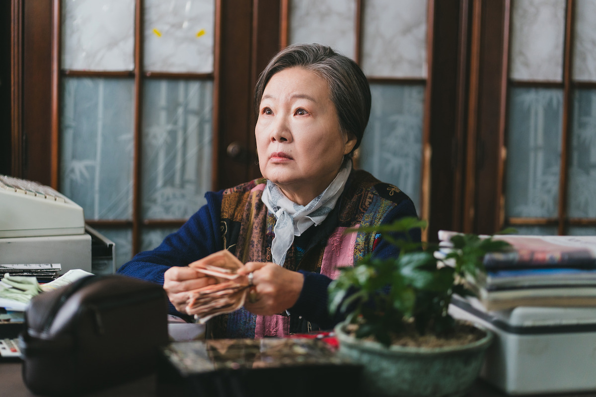 Elderly woman sits at a desk counting money, surrounded by office supplies, a calculator, papers, and a potted plant, in a traditional room with wooden and frosted glass panels.