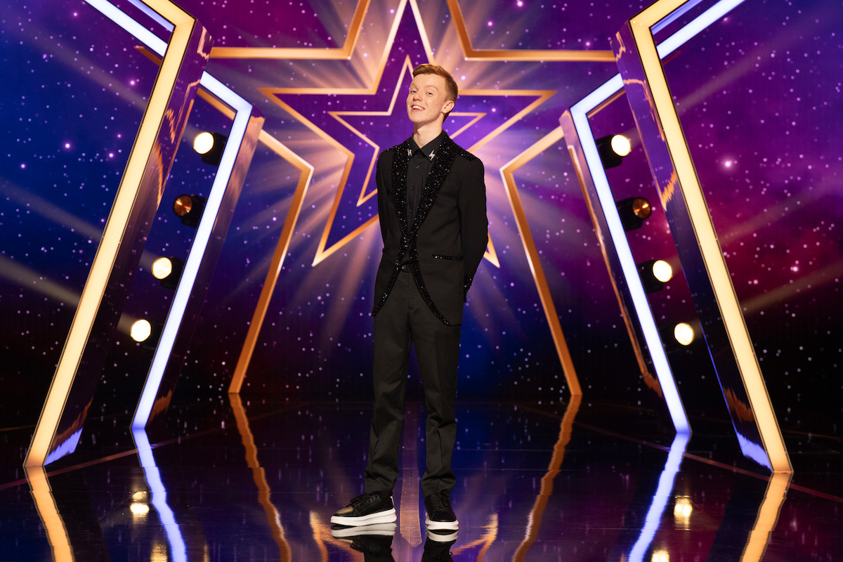 Harry Merlin Piper in a black suit stands on a glossy stage with a large illuminated star, surrounded by purple lights and decorative light bars, creating a vibrant, show-like atmosphere.