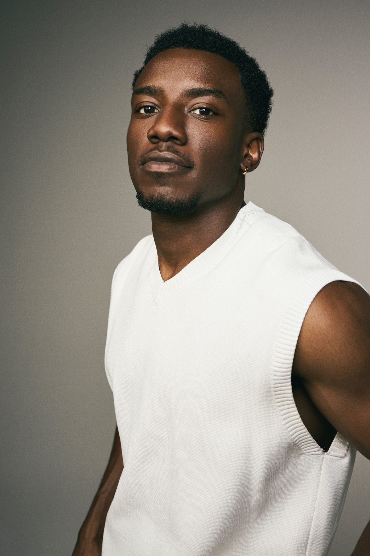 Man wearing a sleeveless white sweater, standing against a plain, neutral background, looking confidently at the camera under soft studio lighting.