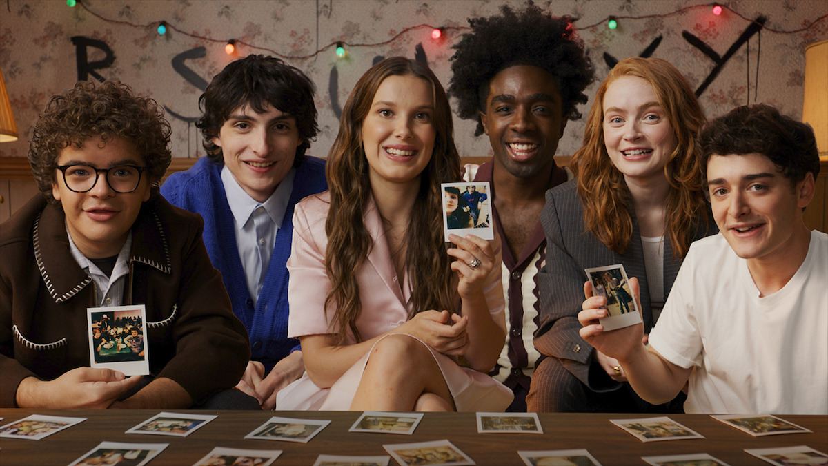 The cast of 'Stranger Things' sits together indoors at a table covered with instant photos, colorful string lights hang behind them, and two of them display photos to the camera, creating a joyful and friendly atmosphere.
