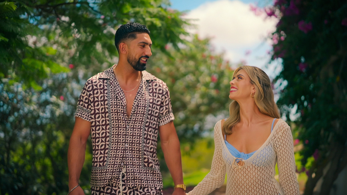 A smiling man and woman holding hands, walking outdoors in a lush garden with green trees and vibrant flowers under a blue sky, wearing summery clothes and looking at each other happily.