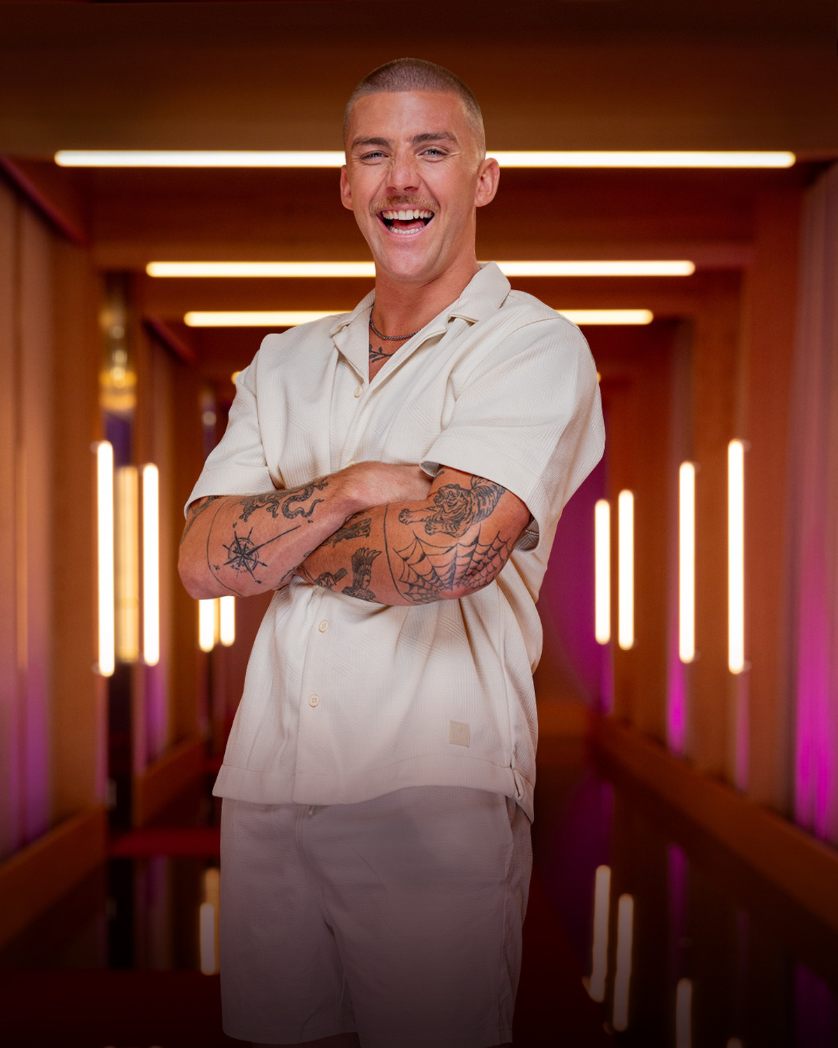 Smiling man with short hair and tattoos, dressed in a light short-sleeve shirt and shorts, standing confidently with arms crossed in a modern, warmly lit hallway with vertical lights and purple accents.