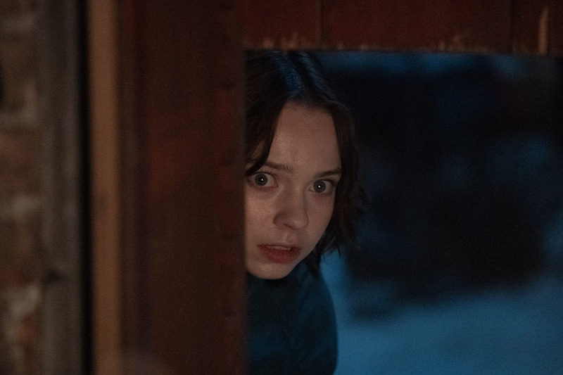 A person with short hair cautiously peeks out from behind a wooden door in a dimly lit, possibly outdoor setting, appearing alert or concerned.