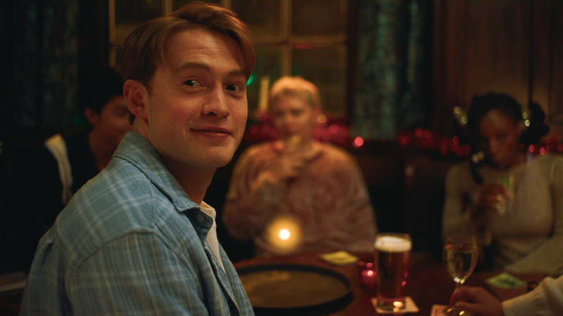 A young man in a blue shirt smiles while sitting at a cozy pub table with friends, drinks, and soft lighting, suggesting a relaxed social gathering in a warm, festive environment.