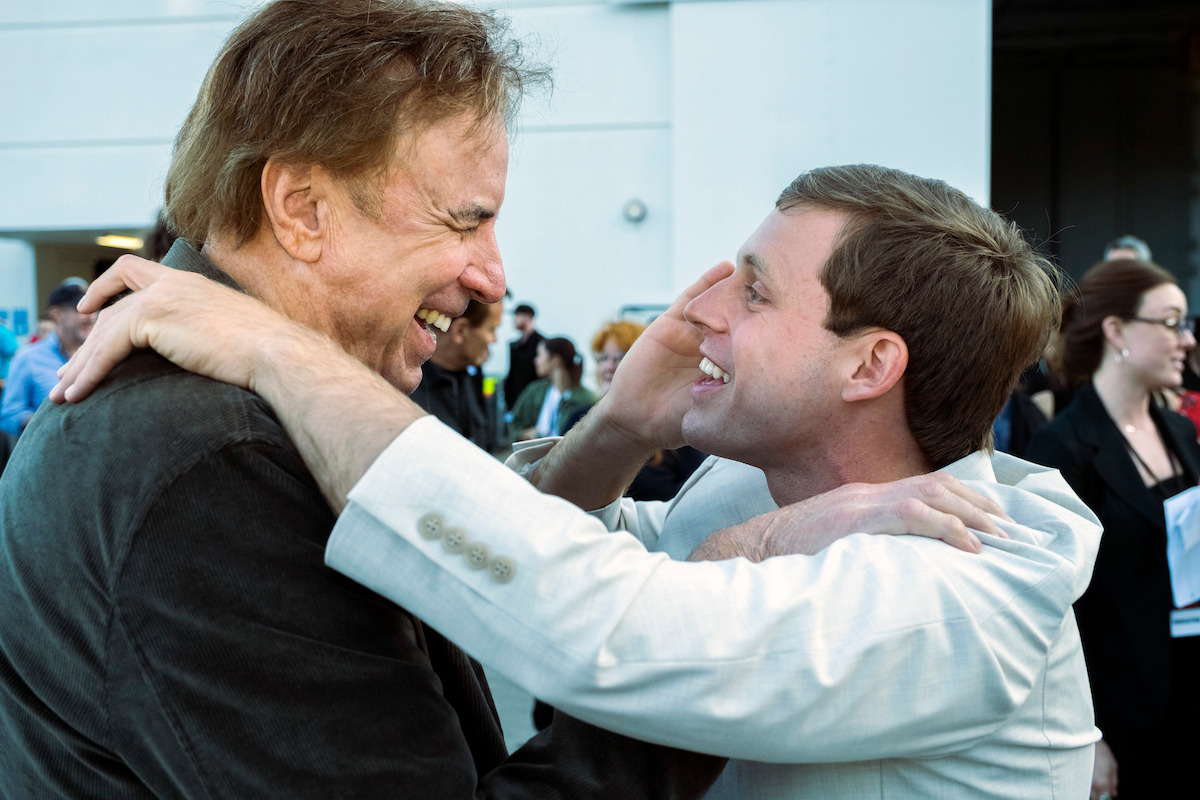 Kevin Nealon and Tanner Smith smiling and embracing outdoors in a cheerful greeting, with people and a building visible in the background, suggesting a social or professional gathering.