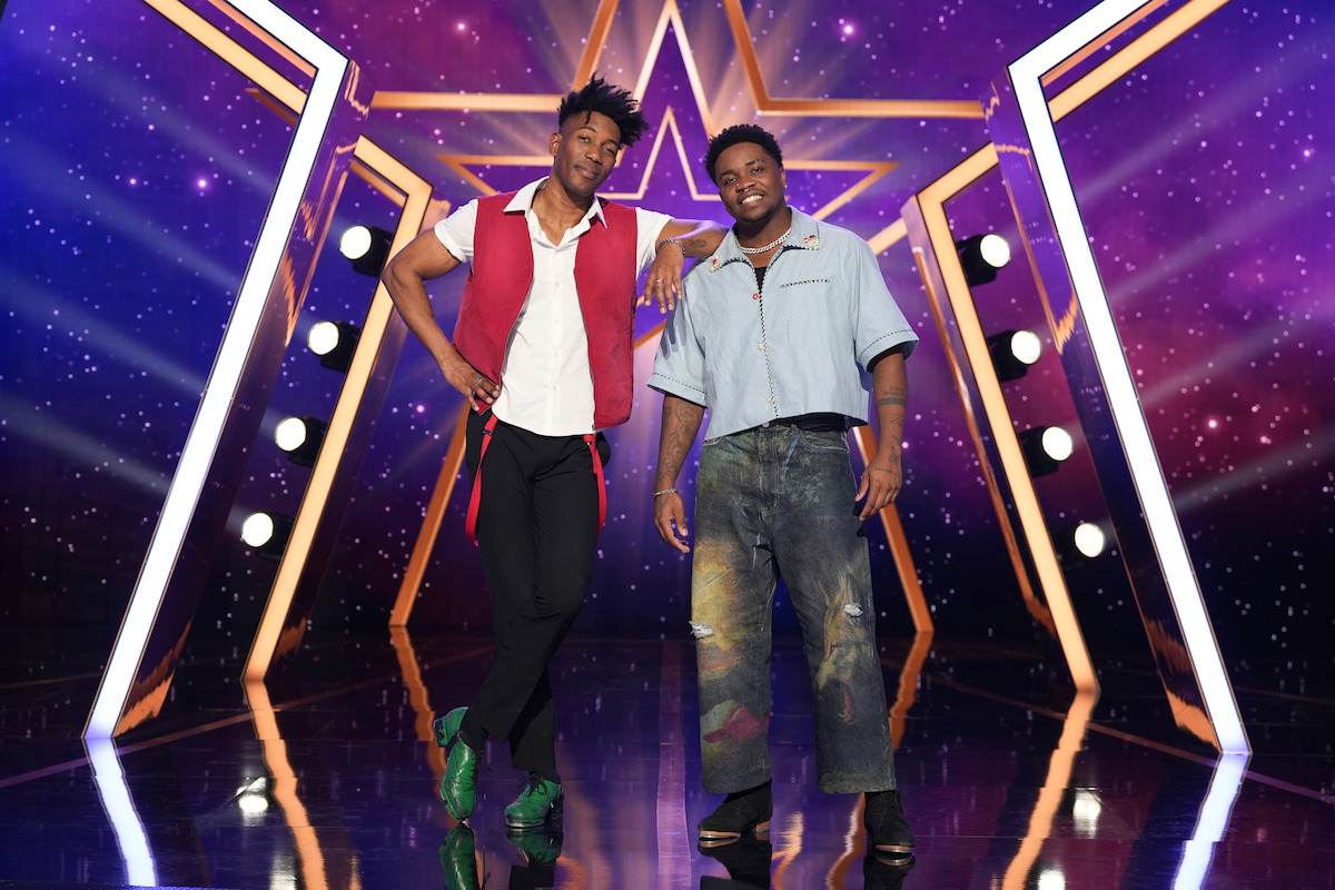 Kenji and Jabu pose smiling on a stage with a glowing star backdrop, surrounded by stage lights and a purple, starry sky-themed environment, creating a lively and vibrant atmosphere.