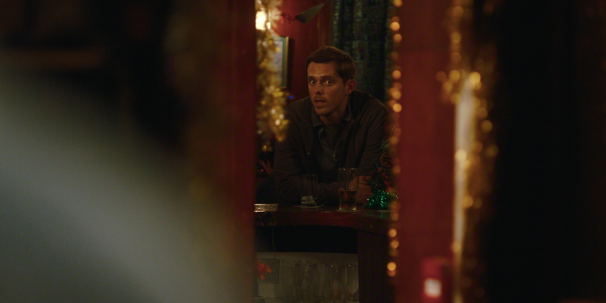 A man leans on a dimly lit bar counter decorated with gold tinsel and festive ornaments, surrounded by glasses and warm ambient lighting, creating a cozy, intimate setting.