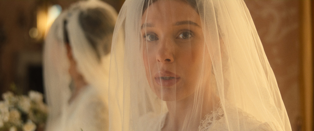 A bride with a white veil and lace dress stands indoors, looking ahead with a soft expression. A mirror reflects her image, and flowers are visible in the background, creating an elegant, warm atmosphere.