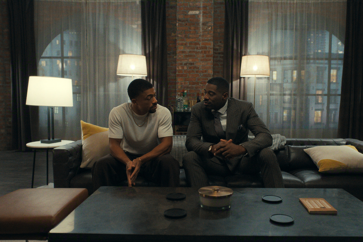 Two men sitting on a couch in a stylish, urban living room at night, engaged in a serious conversation with city lights visible through large windows and warm lamps illuminating the space.