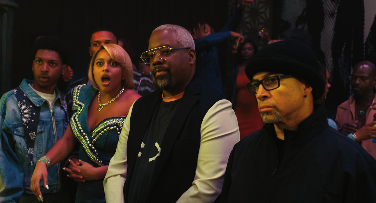 A group of people at a lively indoor event with colorful lights, one woman in a sparkling dress looks surprised, while two men in glasses stand in the foreground, one wearing a beanie, both appearing serious.