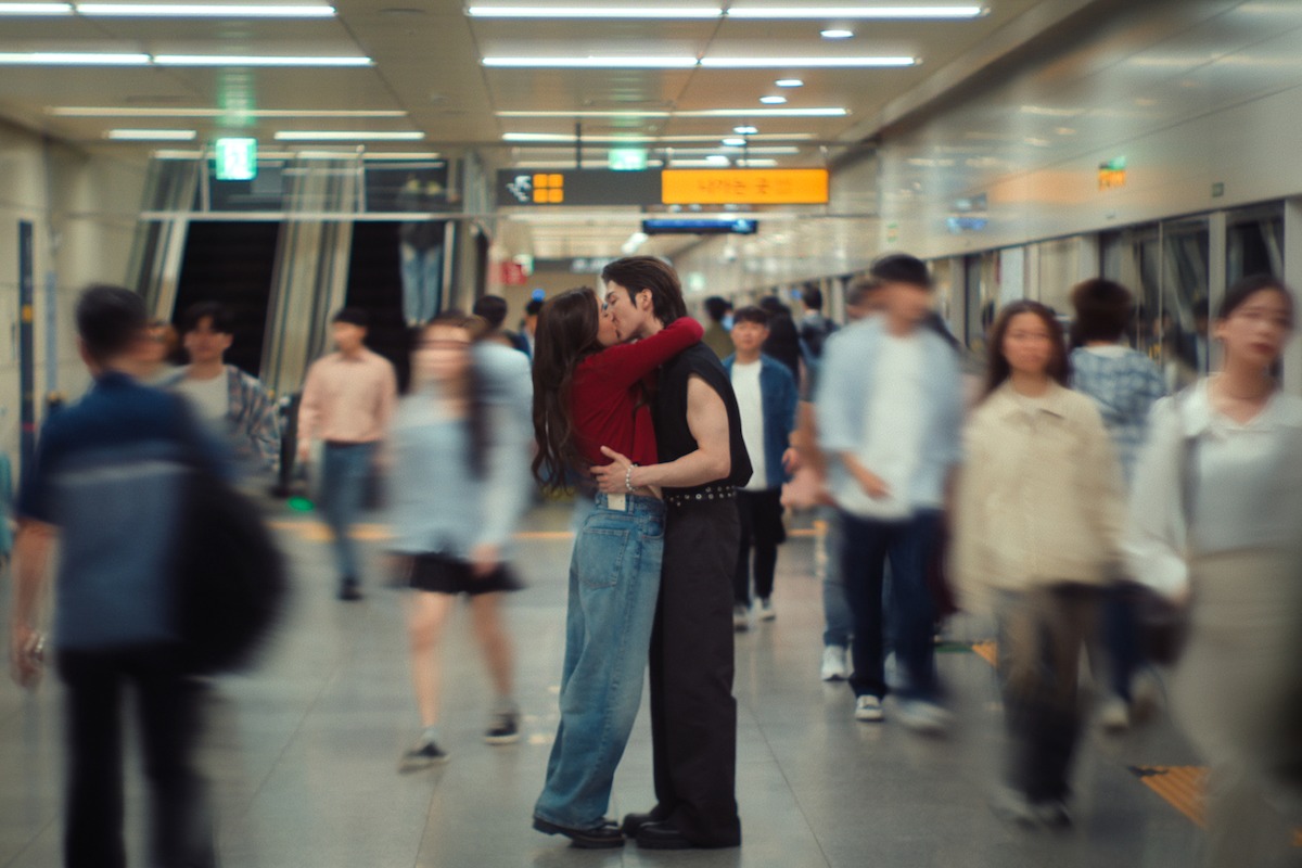 A couple embracing and kissing in the middle of a busy subway station, surrounded by blurred commuters walking past them under bright fluorescent lights.