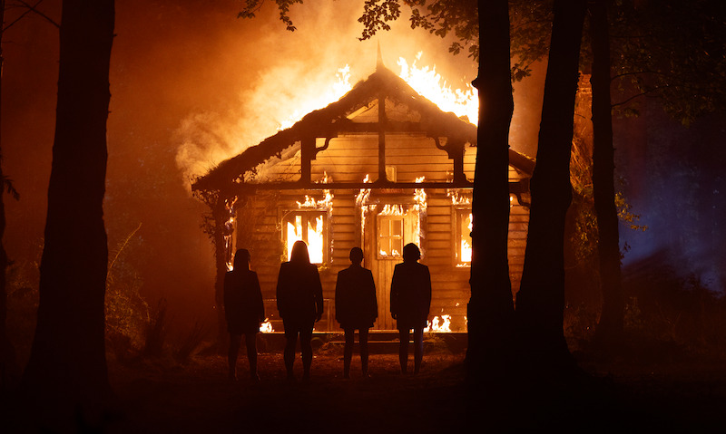 Four people stand silhouetted in a dark forest, watching a wooden cabin engulfed in flames at night, creating a tense and dramatic atmosphere.