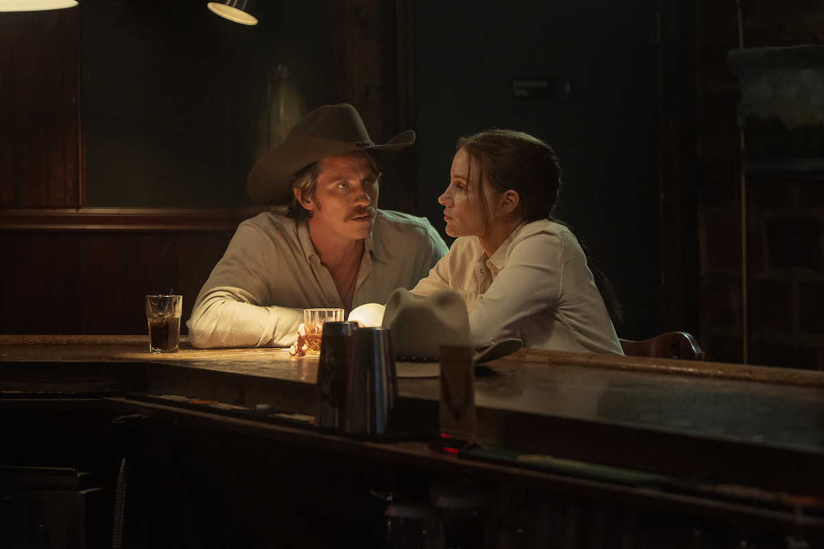 A man in a cowboy hat and a woman sit closely at a dimly lit bar, talking seriously. Glasses of drink rest on the bar, and the setting is cozy with warm lighting and rustic wood and brick surroundings.
