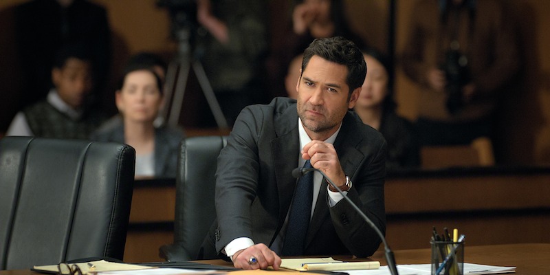 A serious man in a suit sits at a desk in a courtroom, speaking into a microphone. Papers are spread in front of him, with people and cameras in the background, creating a tense, formal atmosphere.
