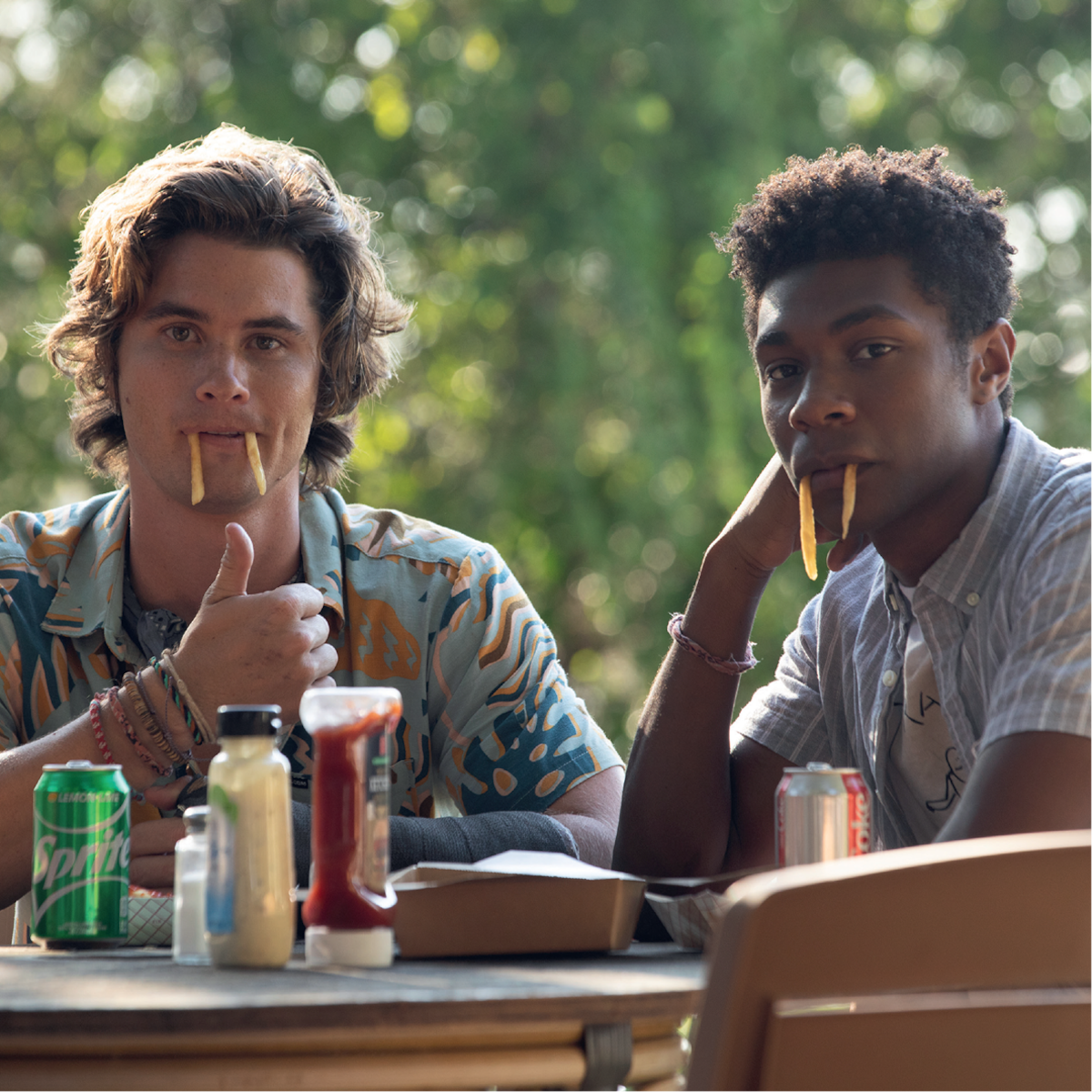 Two young men sitting at an outdoor table, playfully holding French fries in their mouths like fangs, surrounded by soda cans, condiments, and food containers, with greenery in the background.