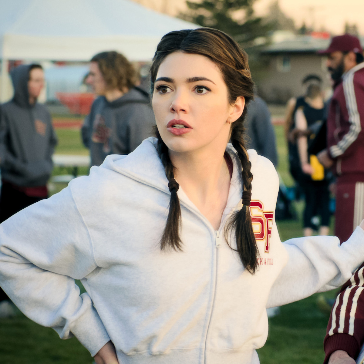 Young woman with braided hair in a gray hoodie stands outdoors at an athletic event, looking focused. Other people in sportswear and tents are visible in the background on a grassy field.
