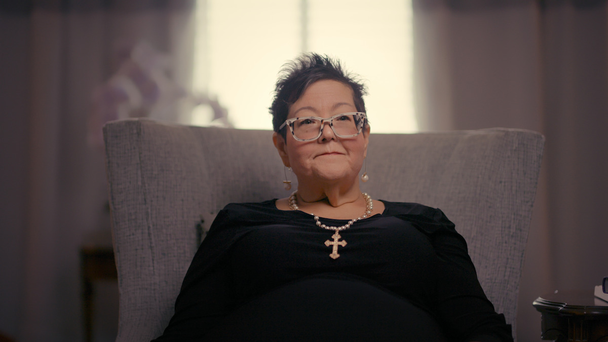 Woman with short dark hair and glasses sits in a gray armchair, wearing a black top and a large cross necklace, with a window and soft curtains in the background of a warmly lit room.