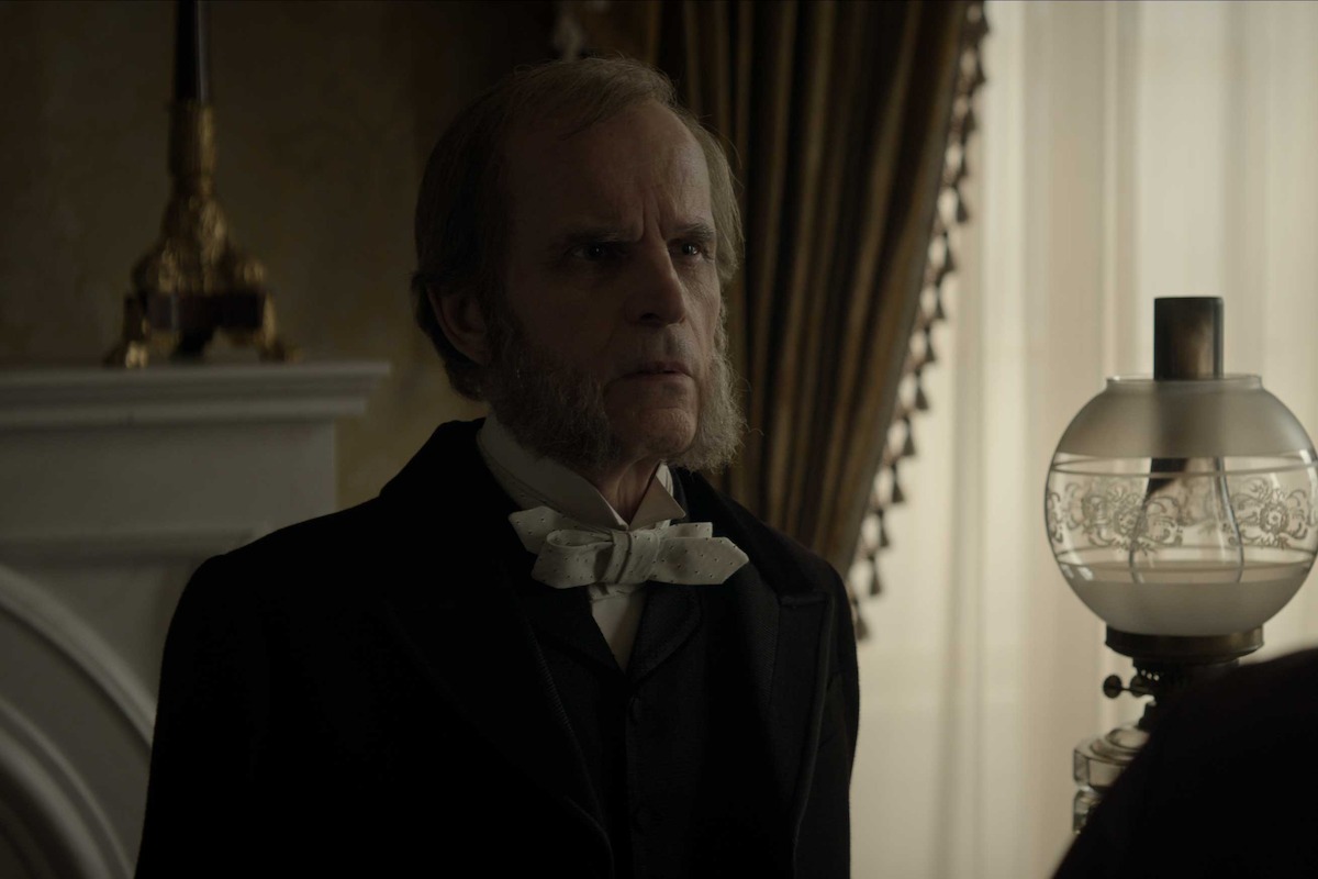 A stern Victorian-era man in formal attire stands in a dimly lit, elegant room with heavy drapes, a white fireplace, and an ornate oil lamp, creating a tense and serious atmosphere.