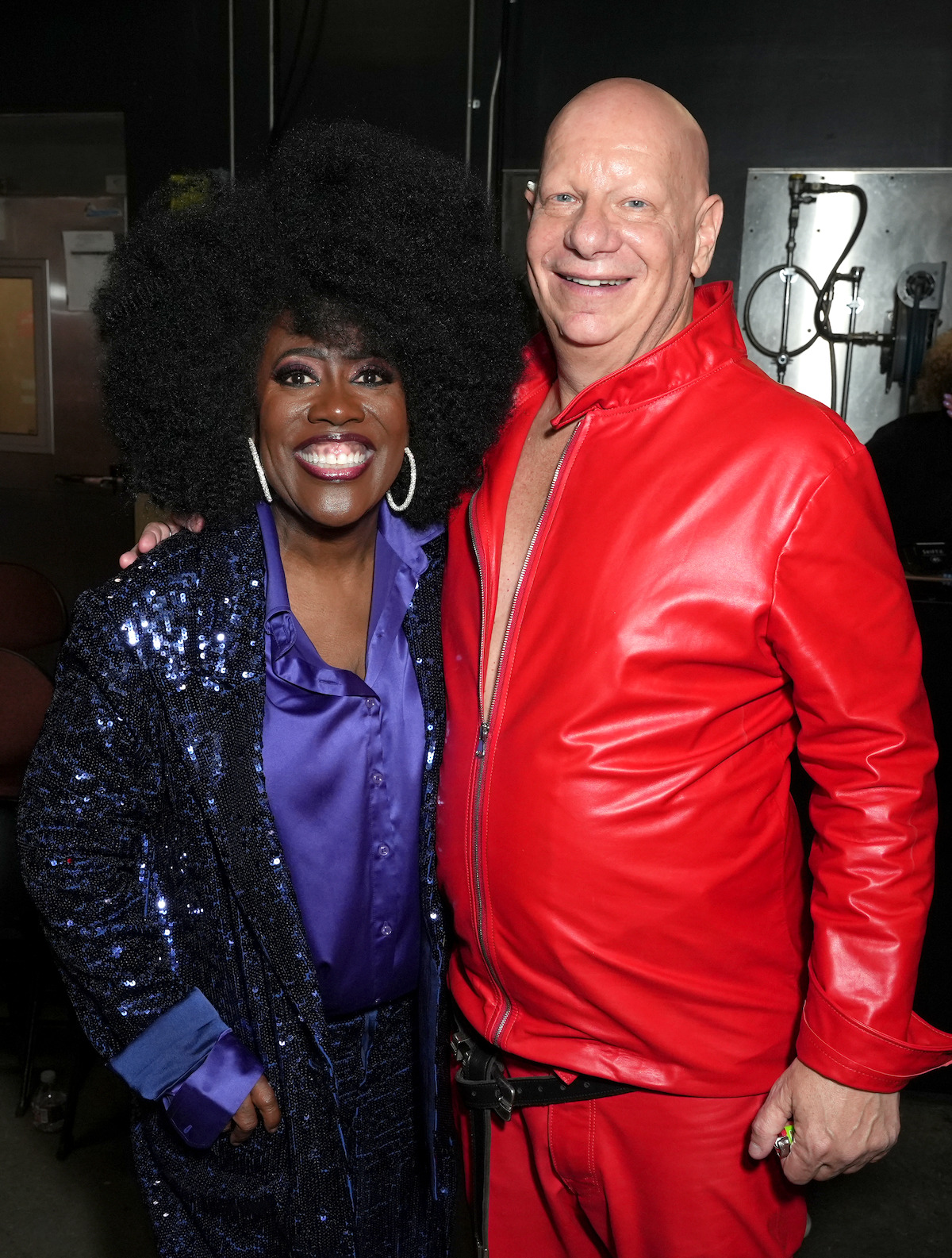 Two people smiling backstage, one wearing a sparkly blue outfit with a purple shirt and large earrings, the other in a bright red leather outfit with a partially unzipped jacket. Industrial setting with dim lighting and visible equipment.