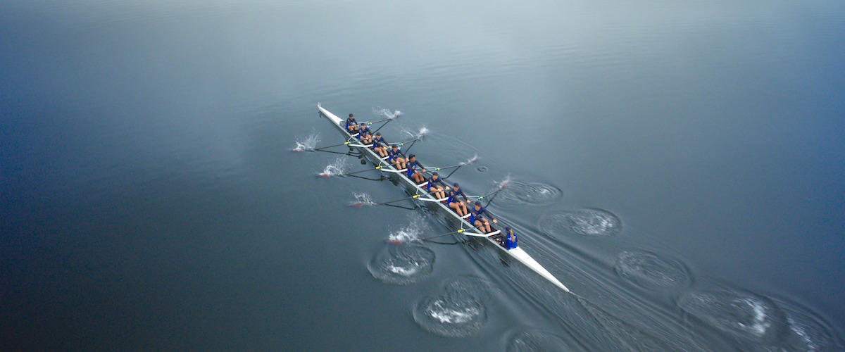 Aerial view of a rowing team in a long boat moving in sync across calm, reflective water, creating ripples as they row together in a misty, tranquil outdoor environment.