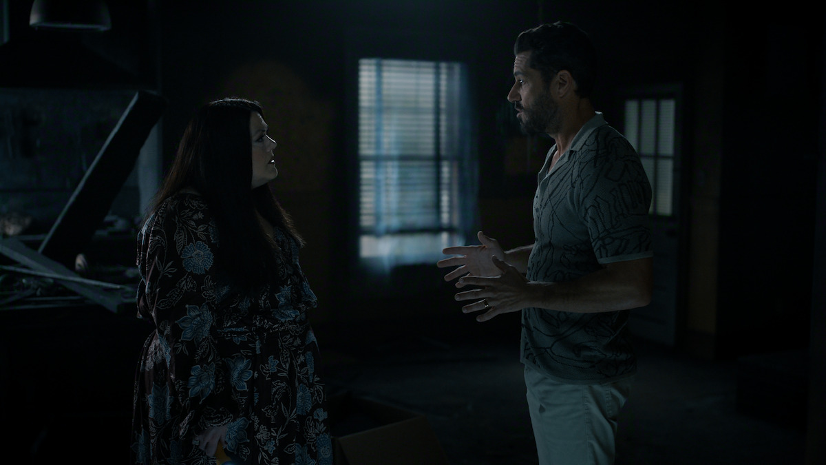 Two people standing and talking in a dimly lit room with closed blinds and scattered furniture, creating a tense atmosphere.