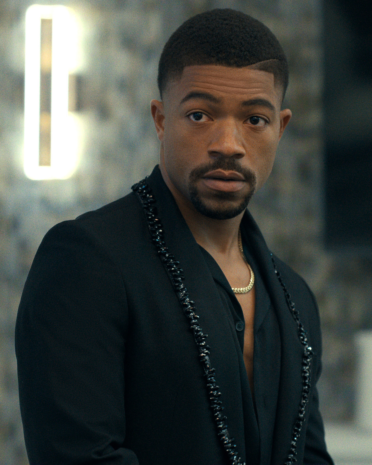 A well-dressed man in a black suit with a chain necklace stands indoors against a blurred modern background with a lit wall fixture, looking slightly to the side.