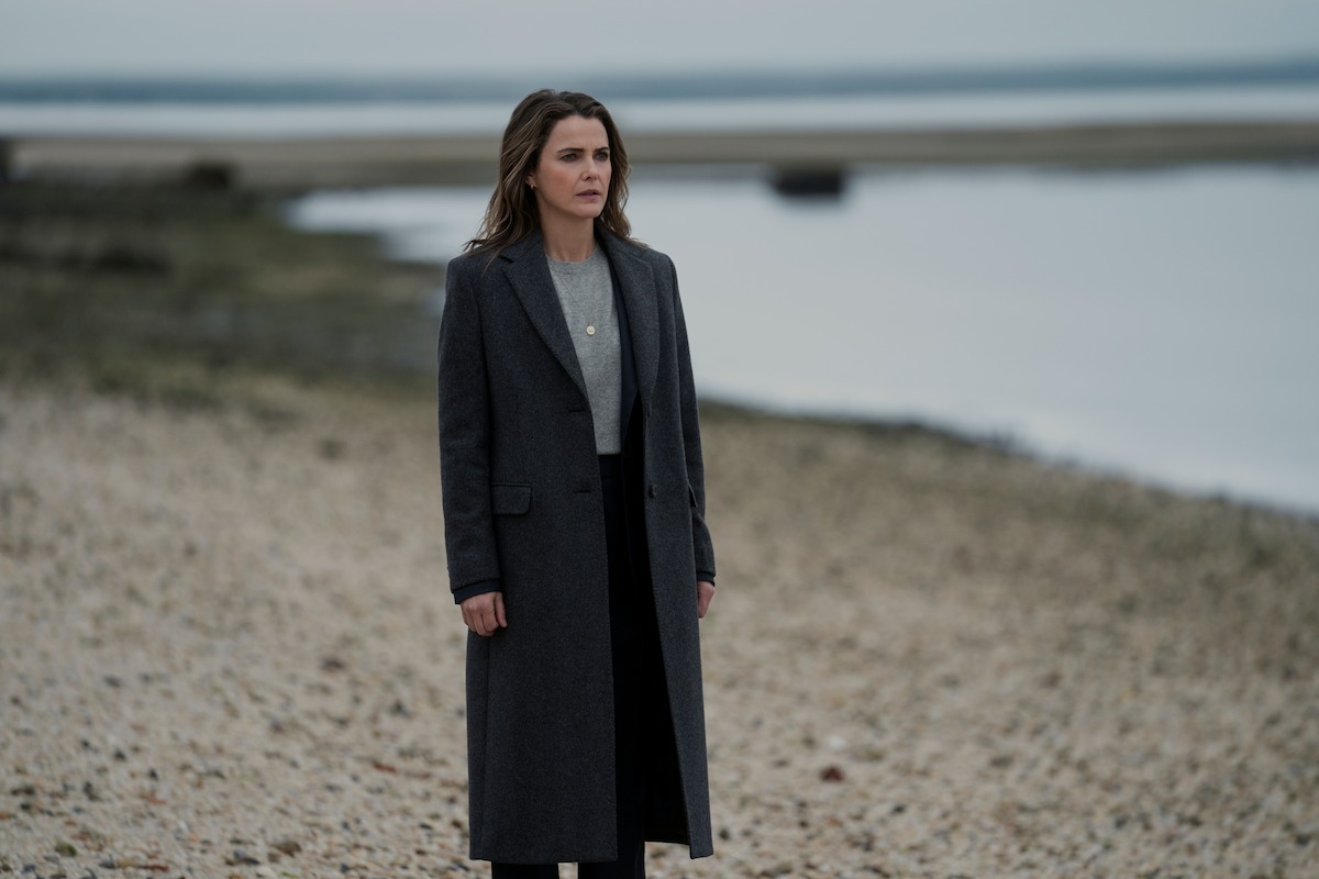 Woman in a long dark coat standing alone on a rocky beach, with calm water and an overcast sky in the background.