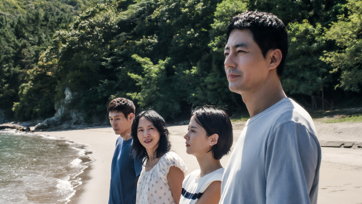 Four people stand side by side on a sandy beach with trees and greenery in the background, looking thoughtful under natural daylight by the shoreline.