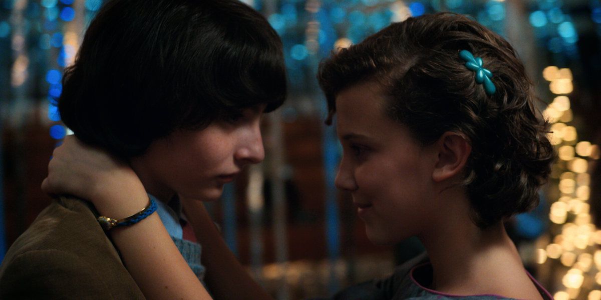 Two teenagers slow-dancing closely at a decorated school dance, surrounded by sparkly blue streamers and warm lights.