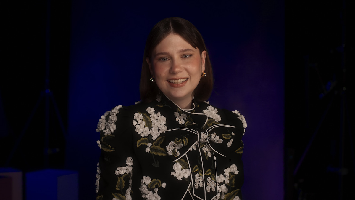 A smiling woman with straight brown hair wears a black floral embroidered jacket, posing indoors against a dark background with subtle purple lighting.