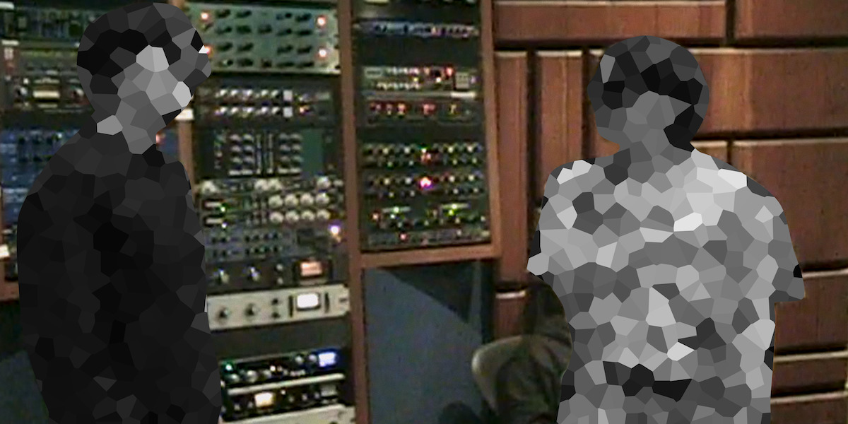 Two people in a recording studio stand near racks of audio equipment and mixers, surrounded by soundproofed walls and acoustic paneling.