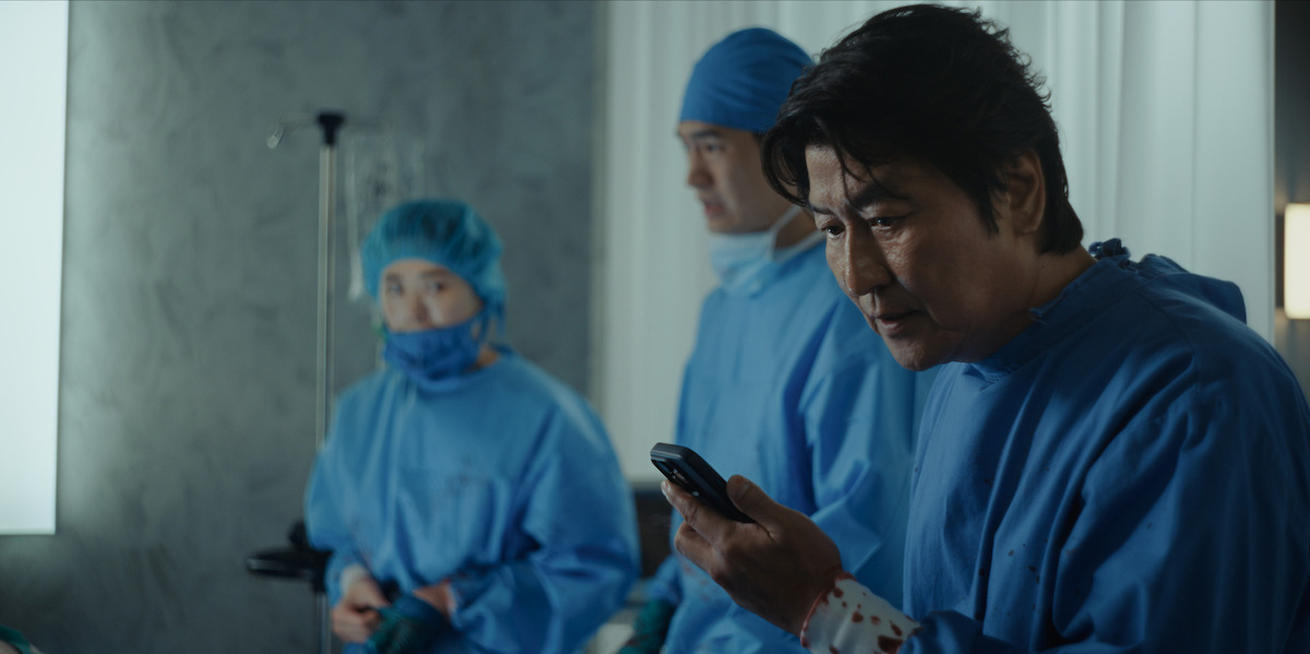 Three people in blue medical scrubs and caps are in a clinical room; one person in front is holding and looking at a phone, while the others stand in the background near an IV stand.
