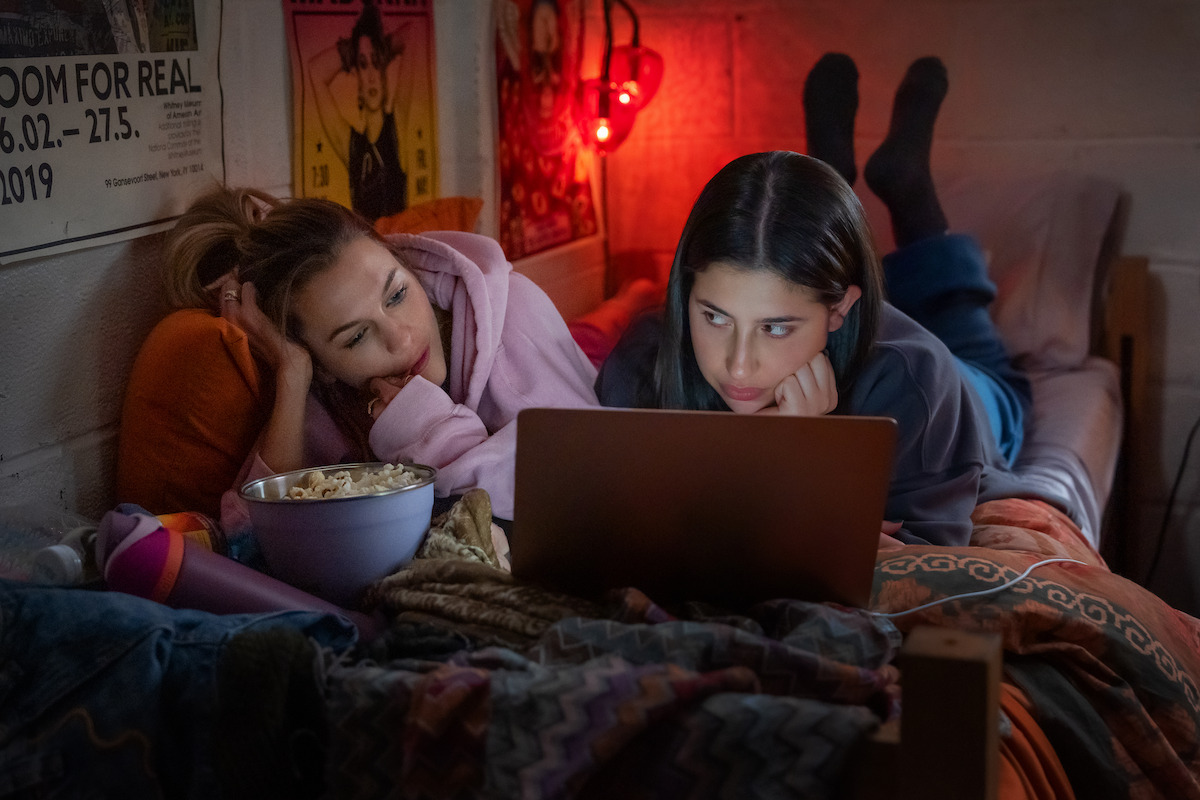 Two young women lying on a bed in a cozy, dimly lit room, watching a laptop together. There is a bowl of popcorn, posters on the wall, and warm blankets, creating a relaxed, intimate atmosphere.
