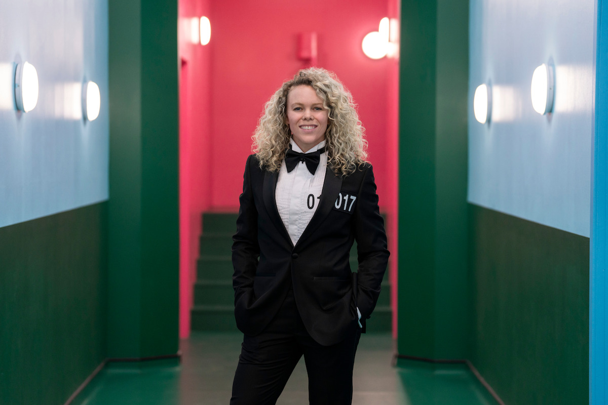 A confident person with curly blonde hair in a tuxedo stands in a brightly lit, colorful hallway with teal walls and a vibrant pink background, creating a modern, energetic atmosphere.