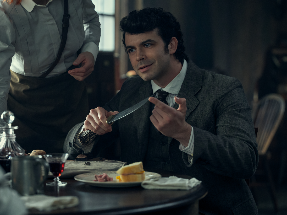 Man in vintage suit sitting at a table in a dimly lit room, holding a knife while talking, with food, drink, and another person in an apron standing nearby, suggesting a historical setting.