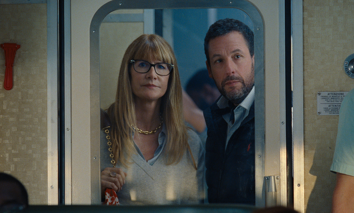Laura Dern with long blonde hair and glasses stands next to a bearded man, Adam Sandler. Both look forward through a glass door on a train.